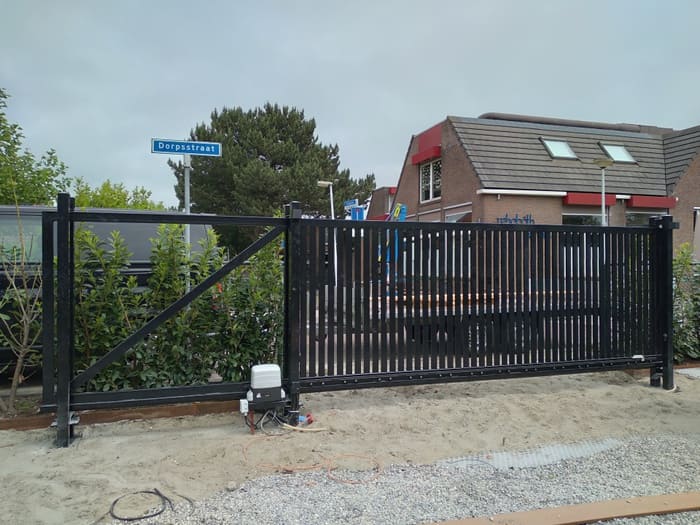 Maatwerk houten schuifpoort met zwart frame en verticale houten latten naast een vaste doorgang bij een woning